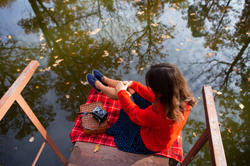 A young woman sits with her back on a bridge near a lake on a sunny day. Plaid, camera, suitcase.