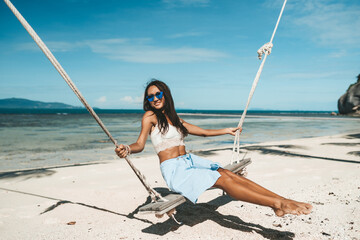 Woman in blue skirt and white top swinging at tropical beach
