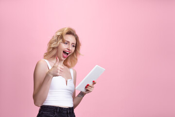 Scrolling tablet, good offer. Caucasian young woman's portrait isolated on pink studio background. Beautiful blonde model. Concept of human emotions, facial expression, sales, ad, youth. Copyspace.