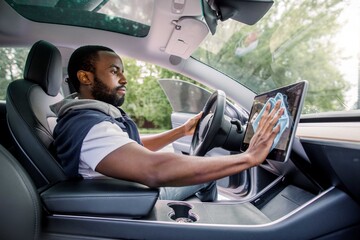 Cleaning time, car detailing concept. Portrait of young handsome African man with beard sitting inside his modern self-steering car and wiping dust from touchscreen display with microfiber cloth