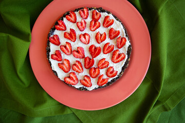 A strawberry cheesecake decorated with heart-shaped strawberries lies on a coral-colored plate and stands on a green napkin. Illustration of a cheesecake recipe with strawberries. View from above.