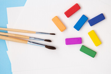 Multi-colored bright paints, watercolor brushes of different sizes lie together on watercolor paper on a blue background. Close-up.