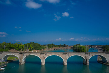 Fototapeta premium Railway arch bridge over the Mincio River, Peschiera del Garda, Italy. Aerial view, Panorama of Lake Garda. Cumulus clouds in the blue sky