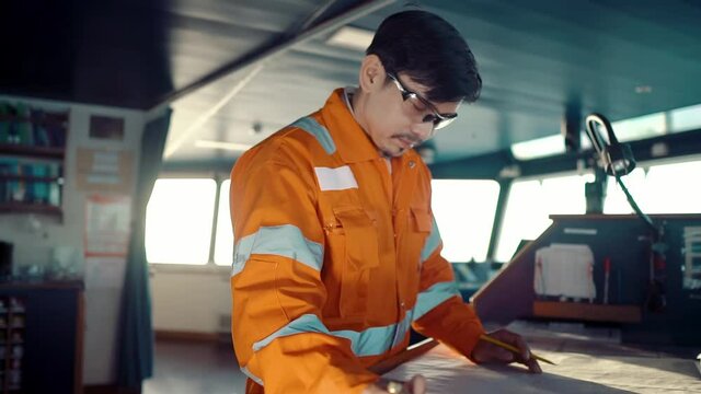 Filipino deck Officer on bridge of vessel or ship wearing coverall during navigaton watch at sea . He is plotting position on chart