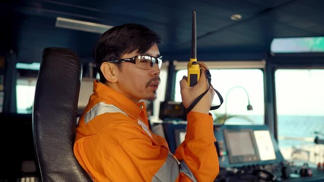 Filipino deck Officer on bridge of vessel or ship wearing coverall during navigaton watch at sea . He is speaking on GMDSS VHF radio, communication between vessels.