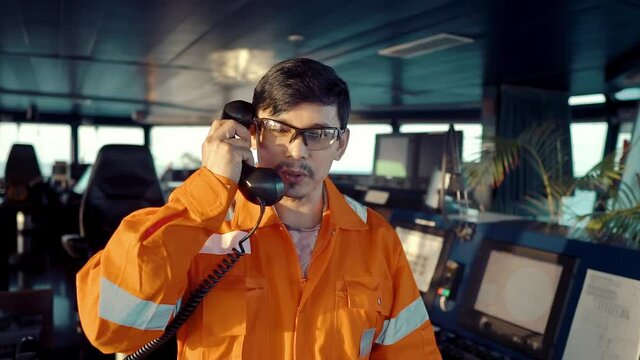 Filipino deck Officer on bridge of vessel or ship wearing coverall during navigaton watch at sea . He is speaking on GMDSS VHF radio, communication between vessels.