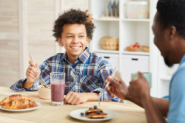 Obraz na płótnie Canvas African boy with curly hair smiling to his father while they having breakfast together in the kitchen