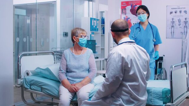 Medical stuff in visor explainingX Ray result to old retired woman sitting on hospital bed during COVID-19 pandemic. Nurse checking with old man in background. Modern private clinic or healthcare