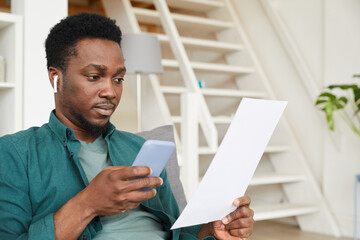 African young businessman sitting on sofa in the room using mobile phone while examining business contract