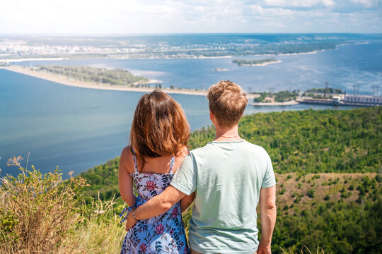 A young couple, a man and a woman, embrace against the background of the Volga, hydroelectric power station and Zhiguli mountains. A photo from the back. Russian tourism.