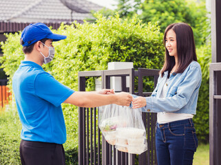 Fototapeta premium Asian young delivery man wear face mask he making grocery service giving rice food boxes plastic bags to woman customer receiving door at house after pandemic coronavirus, Back to new normal concept