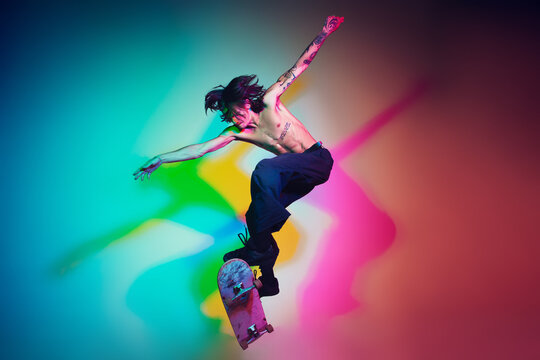 Skateboarder doing a trick isolated on studio background in colorful neon light. Young man shirtless riding and skateboarding in motion. Concept of leisure activity, sport, extreme, hobby and motion.