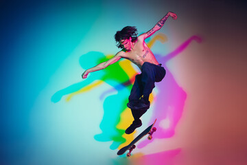 Skateboarder doing a trick isolated on studio background in colorful neon light. Young man shirtless riding and skateboarding in motion. Concept of leisure activity, sport, extreme, hobby and motion.