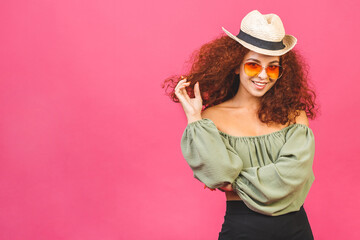 Travel summer concept. Stylish curly young girl or woman in a straw hat with sunglasses isolated over pink background with copy space.