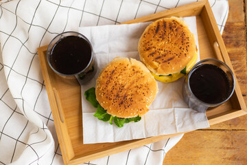 Big and tasty burgers lie on a wooden tray with a glass of cola. Wooden table and white checkered napkin. View from above.