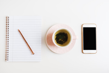 Green tea in a pink cup, mobile phone and notebook with metal pen on white office desk, top view. Freelance concept, flat lay