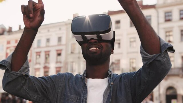 Young African American man playing in virtual reality headset in city center. Guy is using VR glasses and smiling.