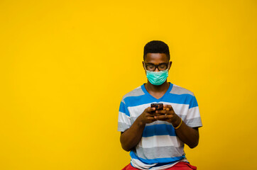 a young handsome african man wearing strip clothe and face mask isolated over yellow background as he prevent himself from the out break in the society and operating his cellphone.