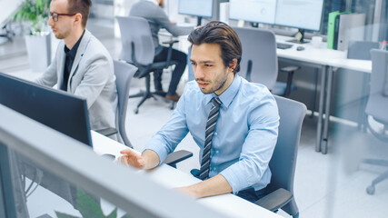 Handsome Motivated Office Worker Uses Desktop Computer, Typing on a Keyboard. Confidentand Young Businessman Works in the Big Bright Office with Team of Professionals