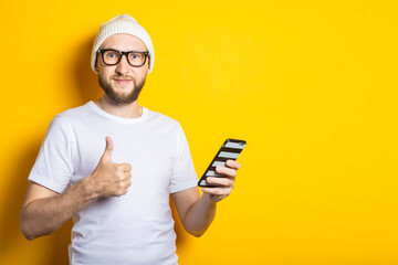 Smiling young man with beard holding phone and holding thumb up class on yellow background.