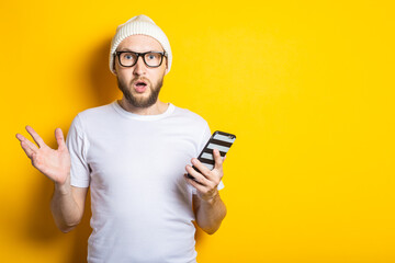 Surprised shocked young man with beard holding phone on yellow background.