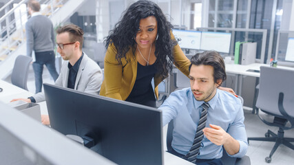 Naklejka premium In Bright Modern Office: Handsome Office Worker Sitting and Working at His Desktop Computer, Discusses Problem Solution with His Female Project Manager. Diverse Team of Businesspeople Solving Problems