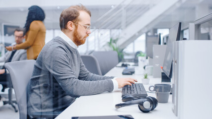 Fototapeta premium Young Happy Business Salesman Specialist Works with His Desktop Computer in Bright Modern Office. In the Background Diverse Team of Professional Businesspeople Working