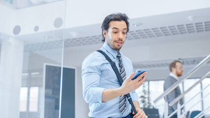 Young Happy Businessman Walks Into Bright Modern Office, Holds Smartphone and Checks Social Media. In the Background Diverse Team of Professional Businesspeople Working