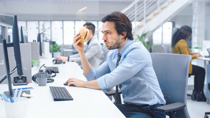 In the Bright Modern Office Young Businessman Eats Hamburger Sitting at His Desktop Computer, He Chewing the Bun and Continues to Work During His Lunch. In the Background Colleagues Working