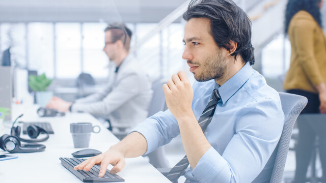 Portrait of Handsome and Confident Shirt and Tie Wearing Businessman Uses Desktop Computer, Typing on a Keyboard, Monitoring Business Transactions, Signing Contracts. Works in the Big Bright Office