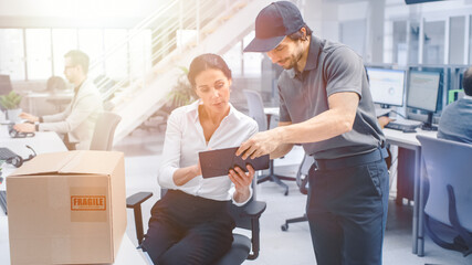 Fototapeta premium In Corporate Office Beautiful Businesswoman Recieves Postal Package from Delivery Man and Signs on Tablet Computer. Modern Office. Shot with Warm Sun Flare Filter.