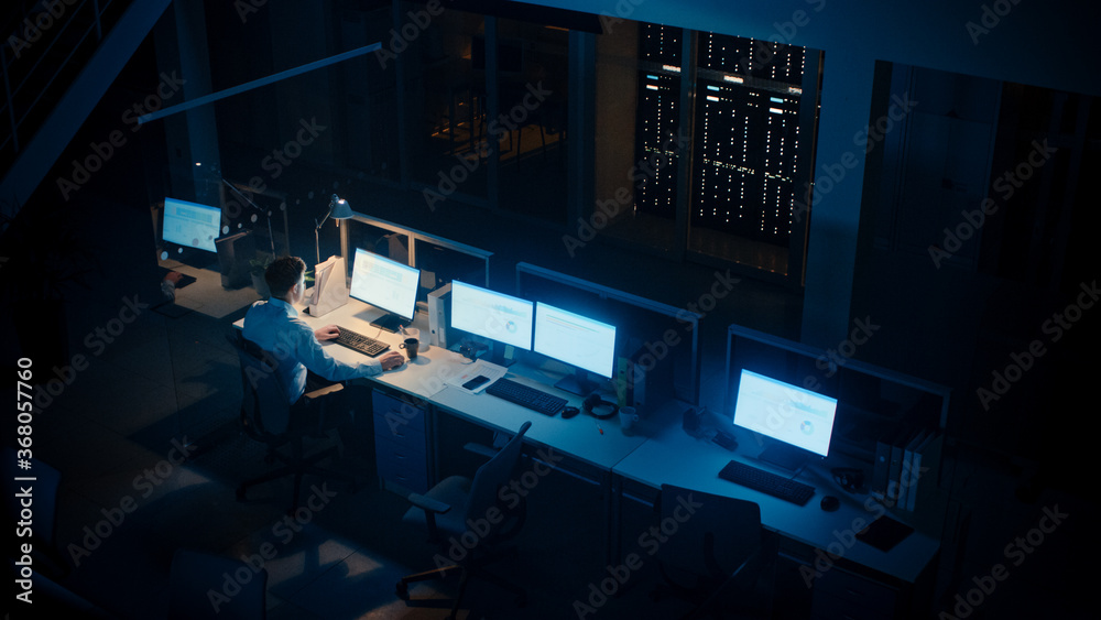 Poster working late at night in the office: businessman using desktop computer, analyzing, using documents,