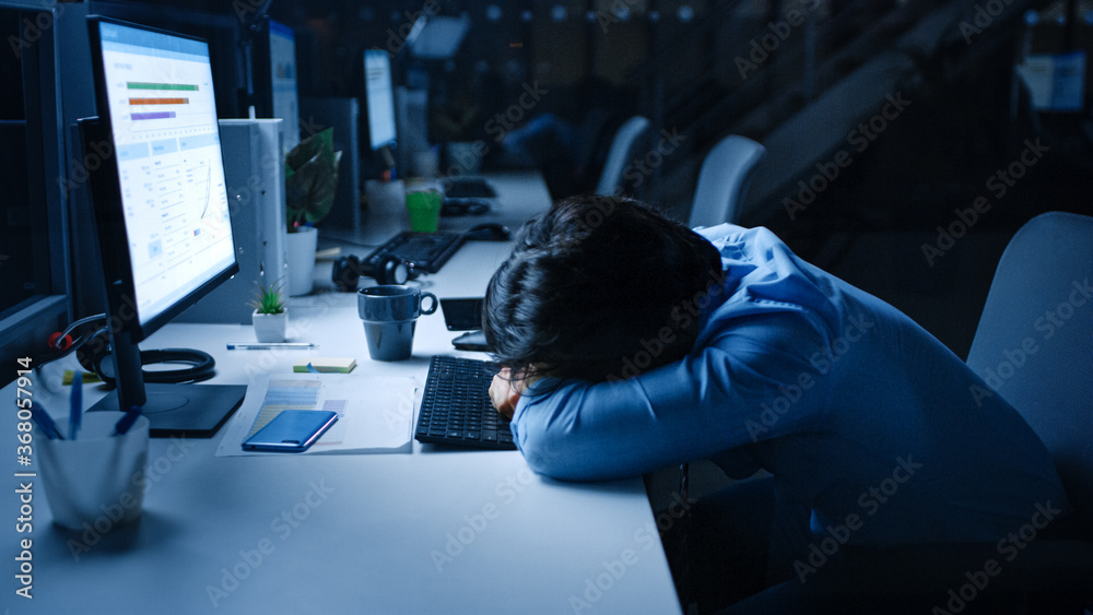 Poster in the office at night overworked tired office worker uses desktop computer but falls asleep fast. t
