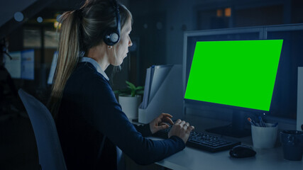Fototapeta na wymiar Working Late at Night in the Office: Handsome Businesswoman with Headphones Using Desktop Compute With Green Mock-up Screen. Call Center Worker, Financial Manager, Emergency Service Worker.