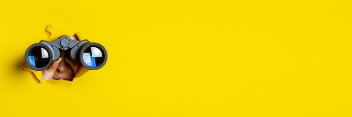 Female hand holds black binoculars on a yellow background. Journey, find and search concept. Banner. - obrazy, fototapety, plakaty