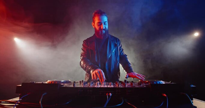 Hipster bearded dj performing in a nightclub. Disc jockey composing a dance music mix at neon light and smoke - nightlife concept 4k footage