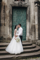 Beautiful wedding couple near the green ancient stone walls. An elegant bride in a satin lace dress smiles with her eyes closed.