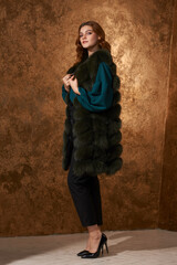Fototapeta premium Stunning young woman wearing blouse, trousers and beautiful fur coat. Luxury, rich lifestyle. Fashion studio shot. alm girl standing against golden wall.