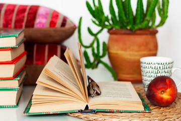 Cozy home interior decor: cup of coffee, stack of books, plants in pots, open book and red peach on a wicker stand, pillows on a white table. Distance home education.Quarantine concept of stay home.