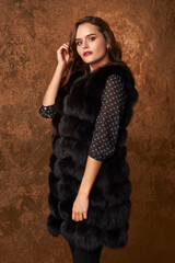 Glamour portrait of beautiful female model with makeup and long wavy brown hair in luxury black fur coat . Studio fashion shot against golden wall.