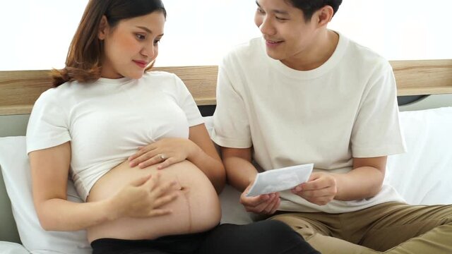 young Asian pregnant woman sits on white bed with husband looking at baby ultrasound image result from hospital