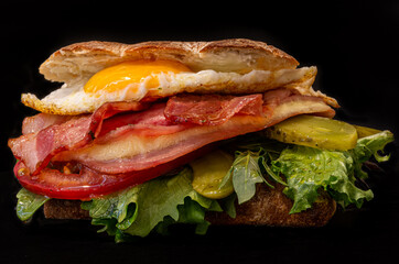 Primer plano de hamburguesa / sandwich con lechuga , tomate, huevo frito, bacon, jamon y queso, sobre fondo negro con patatas fritas.
