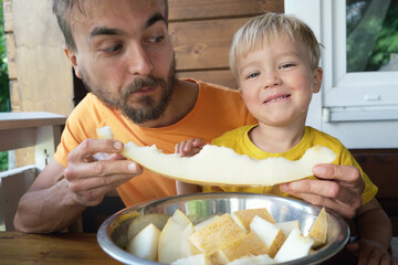 Family eating fresh sweet melon on terrace. Father and his toddler son biting together slice of...