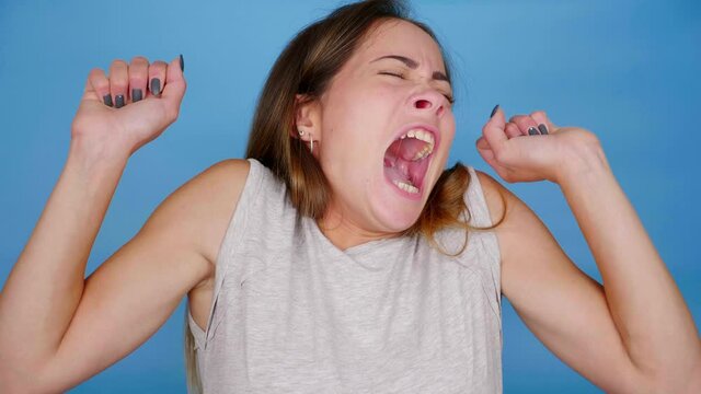 Tired woman in gray t-shirt yawns and stretches on blue background with copy space. 4K slow motion footage