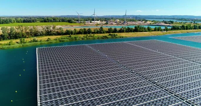 Drone view of solar panels floating in water with windmills, pond and rural aera in the background, under the blue sky. Perfect green energy for a clean environment - 4K