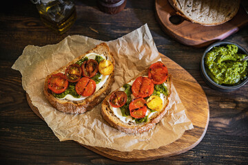 Bruschetta with cream cheese, pesto sauce and grilled cherry tomatoes, herbs on chopping board, rustic wooden table at home kitchen. Italian food, antipasto snack. Selective focus, copy space.