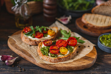 Bruschetta with cream cheese, pesto sauce and grilled cherry tomatoes, herbs on chopping board, rustic wooden table at home kitchen. Italian food, antipasto snack. Selective focus, copy space.
