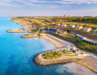 Aerial view of beautiful sandy beach, blue sea, promenade, houses, transparent water, green trees on the hills at sunset in summer. Top view from drone of seafront and cottage town. Tropical landscape