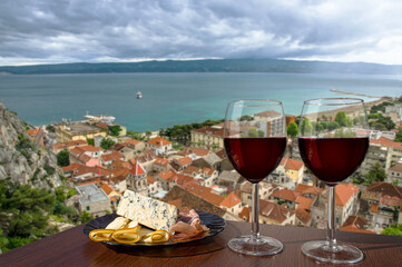 Two glasses of red wine with charcuterie assortment with view of Croatian town Omis with red roofs and blue sea. Glasses of red wine with different snacks - plate with ham, sliced, blue cheese.