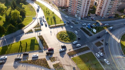 Aerial view of the junction road. There is a roundabout in the middle. Multi lane road, trees and buildings can be seen. Vehicles are waiting in the red light. There is summer and sunset light.